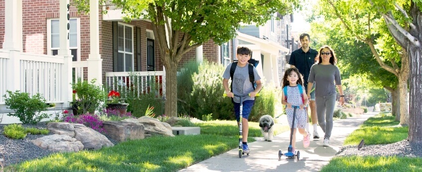 A young family with their small dog walking on a tree lined sidewalk in front of homes, the two kids are on scooters